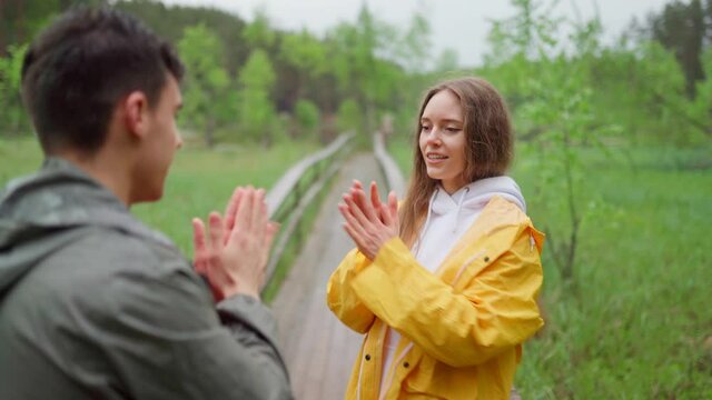 Joyful young couple of hikers having fun playing clap game in nature. Cheerful man and woman in raincoats spending time in countryside on rainy day