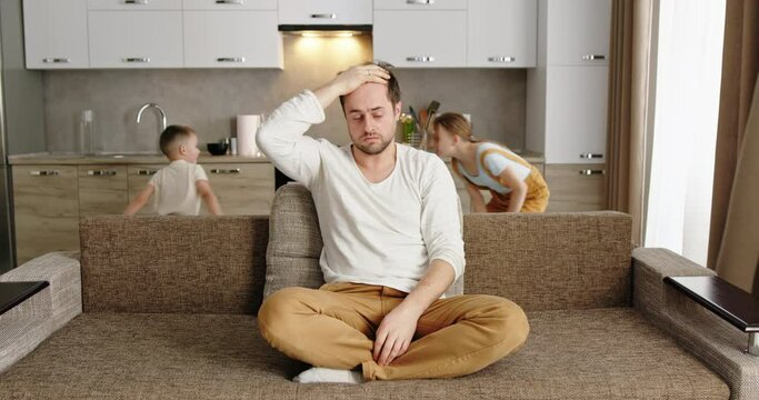 Father sitting on sofa at home exhausted frustrated tired holding his head headache 2 children running around the sofa shouting throwing pillows