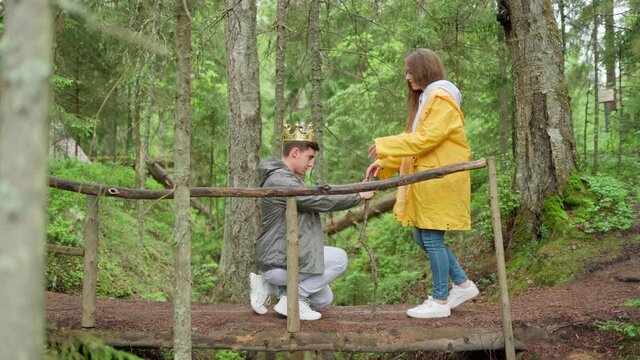 Playful young couple of hikers having fun on wooden bridge over creek in forest. Woman coronating her kneeling boyfriend, putting crown on his head and giving accolade with stick. Happy lovers hugging