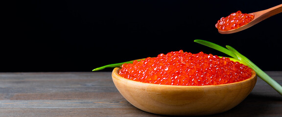 Red caviar in a wooden cup on a black background with a spoon and green onions. A large pile of bright caviar. Fresh delicious caviar. A place for advertising, logo, label, layout, mockup.