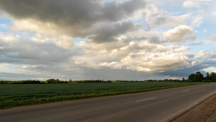 Clouds at sunset over an asphalted empty highway that runs between rural fields. Summer time. Horizontal photo. 
