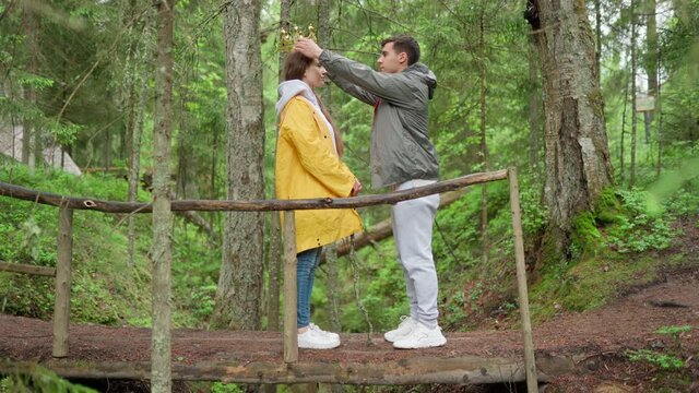 Playful young couple of hikers having fun on wooden bridge over creek in forest. Guy coronating his girlfriend, putting crown on her head and giving accolade with stick. Happy lovers embracing