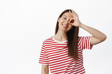 Obraz na płótnie Canvas Portrait of young happy girl shows okay, zero OK sign near eye and smiling, tilt head carefree, positive and joyful pose, wearing summer t-shirt, white background