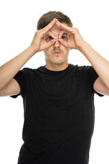 Fototapeta premium Young handsome tall slim white man with brown hair using his hands like a binoculars in black shirt isolated on white background