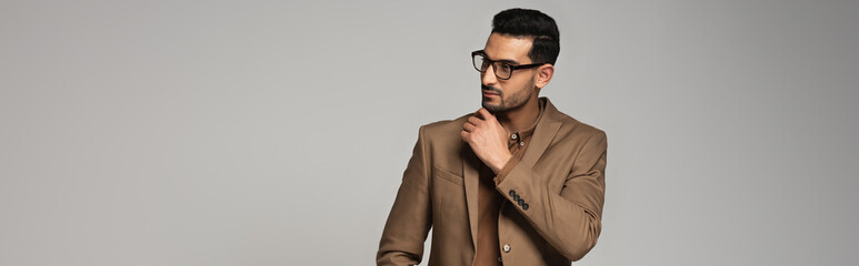 Arabian businessman in eyeglasses and brown jacket looking away isolated on grey, banner