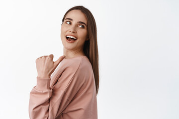 Fototapeta premium Portrait of cheerful beautiful girl pointing thumb and look behind her shoulder, laughing and smiling while looking and showing advertisement logo, white background