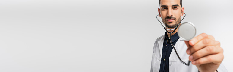 Arabian doctor holding stethoscope on blurred foreground isolated on grey, banner
