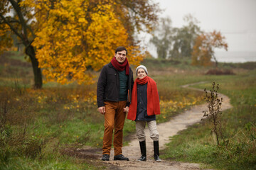 Fototapeta premium Autumn walk outdoors. Dad and pre-teen son in autumn park. Family dressed up in autumn-style clothes (coats, orange scarves, hats) walk in autumn landscape. Alley covered with yellow maple foliage.