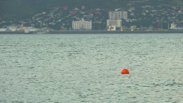 Orange buoy on the water. Ripples in the water. Water marking. Restriction of movement. Signal float. Mountains on the shore. Settlement in the background