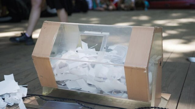 The self-made box from transparent plastic is glued in form of drum for hoax. People throw white sheets of paper with lottery numbers through open hole. Prize draw. Drum for raffle on stage