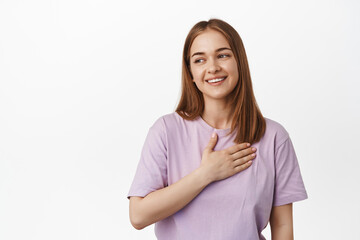 Portrait of beautiful young woman holds hand on heart, looking away at logo banner with pleased, flattered smile, laughing with genuine face expression, stands in t-shirt against white background