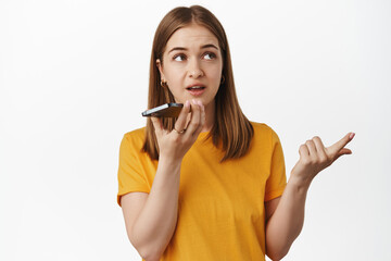 Woman counting while on speakerphone, dictating on mobile phone, thinking and naming smth, record voice message, standing in yellow t-shirt over white background