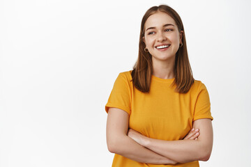 Obraz premium Beautiful young woman holds arms crossed on chest, laughing and smiling, looking aside at promotional text on left empty space, advertising, standing in yellow t-shirt against white background