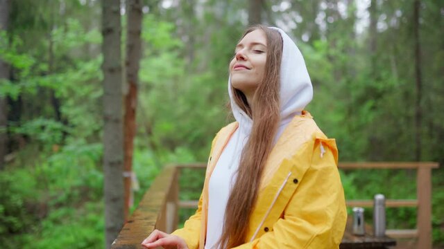 Zoom in of female traveler in yellow raincoat and hoodie breathing deeply and closing eyes enjoying fresh forest air and smell of rain standing on wooden balcony in summer morning