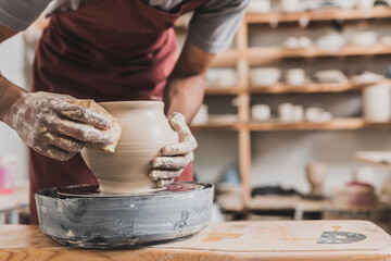 partial view of young african american man making wet clay pot on wheel with sponge in pottery