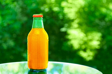 Orange juice in a glass bottle on a mirrored tabletop in the summer outdoors against  background of green foliage.