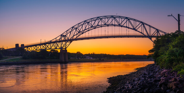 Sagamore Bridge Sunrise over Cape Cod Canal. Dawn Silhouette Architectural Image with Orange and Golden Colors. High Contrast Panoramic Seascape Image with Space for Text and Design.