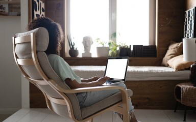Fototapeta premium Mixed race Woman Working On Laptop Computer With Blank White Screen At Home, Sitting In Chair, Over Shoulder View