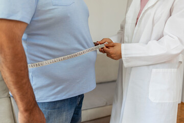 Cropped shot of a female doctor taking obese patient's body fat measurements. Dietitian measuring abdominal circumference during checkup. Young doctor taking measurement from depressed overweight man.