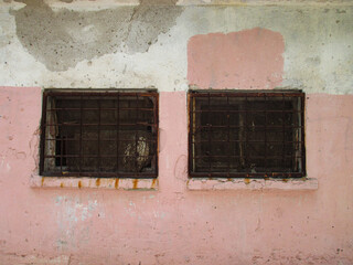 Two windows with metal bars and plates on the facade of the building. Old wall with remnants of pink paint and white stucco. 