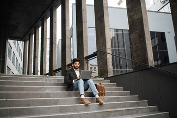 Confident hindu man in stylish clothes working on wireless laptop outdoors. Handsome young freelancer sitting on stairs and looking aside. - obrazy, fototapety, plakaty