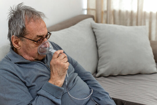 Portrait of elderly man with flu inhalation at home. The senior Caucasian man does inhalation with a nebulizer mask at home for coronavirus disease, sitting on couch at home alone. Health care concept