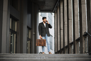 Positive hindu man in stylish formal clothes standing near modern building and talking on cell phone. Smiling businessman with suitcase in hand having mobile conversation outdoors.