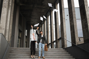 Cheerful arabian man and woman in formal clothes throwing up some documents while standing on stairs outdoors. Concept of common project and successful cooperation.