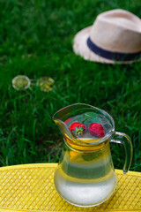 Defocus two strawberry, slice lemon and leaves of mint in glass jug of lemonade on yellow board. Blurred grass background. Pitcher of cool fresh cold drink. Hat and sunglasses. Out of focus