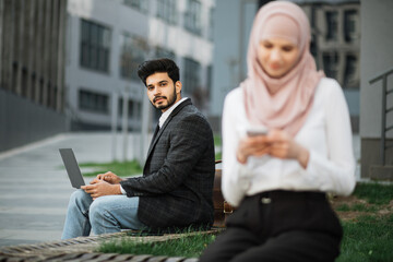 Handsome hindu man sitting on wooden bench and using wireless laptop. Blur foreground of muslim woman sitting near with smartphone in hands. Two coworkers using gadgets outdoors.