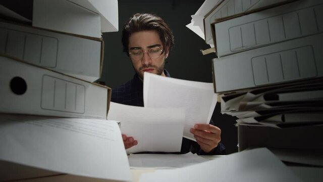 Tired dark-haired man with pile of folders working in office, takes glasses off, employee works with documents at night having headache, exhausted worker holding his head, fatigue, lots of work. 