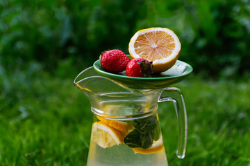 Defocus saucer with lemon and two strawberry standing on glass jug of lemonade with slice lemon and...