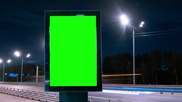 Modern billboard with a green screen on a busy defocused highway with traffic, neon lights. Timelapse of car lights. Blank billboard for advertising. 