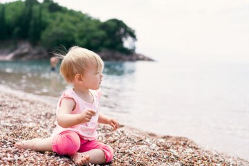 Fototapeta na wymiar Little girl sits on a pebble beach near the water against the backdrop of a green island