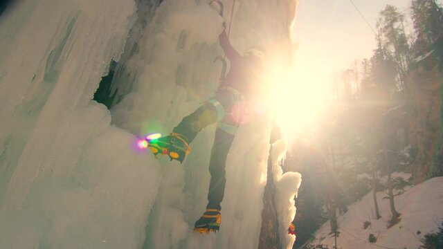 Handheld shot of a female ice climber hiking on a vertical frozen ice surface, making squat, stand, and swinging the ice axes movements