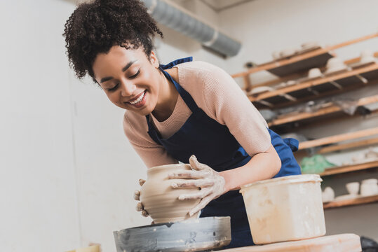 smiling young african american woman shaping wet clay pot on wheel with hands in pottery - Powered by Adobe