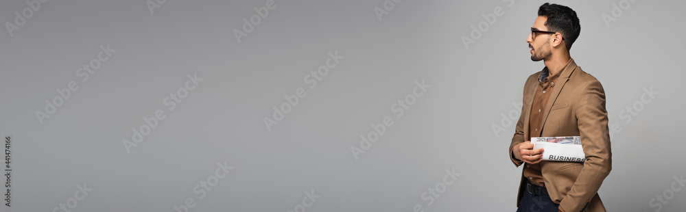 Wall mural side view of muslim businessman with newspaper looking away isolated on grey, banner