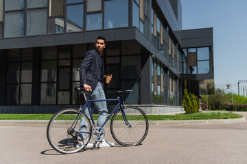 Fototapeta na wymiar Arabian businessman walking near bike and holding paper cup on urban street