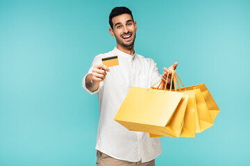 Positive arabian man holding credit card and yellow shopping bags isolated on blue