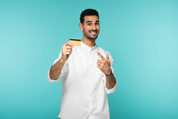 Cheerful arabian man pointing at credit card isolated on blue