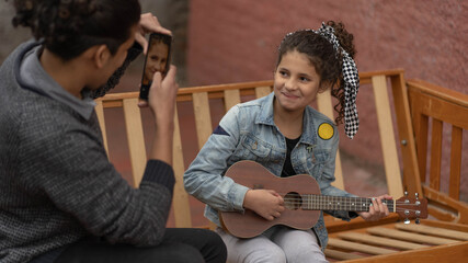 .little girl plays ukulele smiling wearing denim jacket outdoors and transmits stream from...