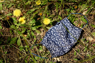 A beautiful blue in floral pattern protective mask lies on the grass in a park among yellow dandelion flowers.