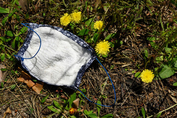 Fototapeta premium Used medical protective face mask lies on the grass in the park among the yellow flowers of the dandelion.