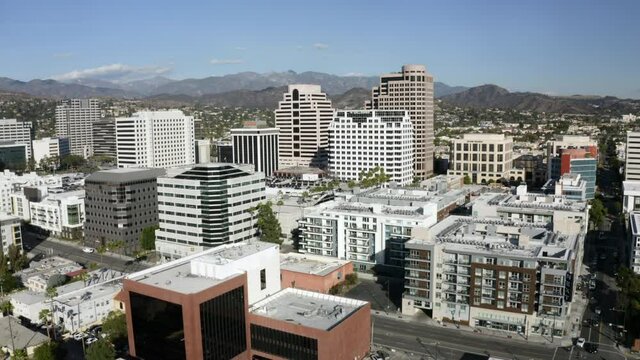 Aerial Tilt-Up To Downtown City Buildings And Urban Skyline With Bright Sunlight, White Towers, And Rugged California Mountains On The Horizon - Glendale, California