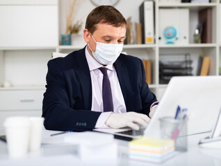 Focused male in disposable personal protective equipment working in business office using laptop, new normal due to coronavirus outbreak