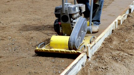 Male worker guides a small yellow compactor on dirt before concrete is poured for driveway, at a new home construction job site.