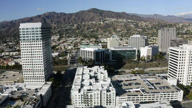 Aerial Panning Bustling Downtown City Buildings And Urban Skyline With Bright Sunlight, White Towers, And Rugged California Mountains On The Horizon - Glendale, California