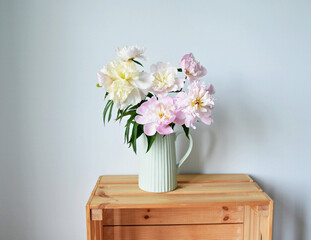 Pink and white peonies in light green ceramic jug on wooden box on white background