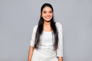 Portrait of happy and positive Asian woman smiling carefree, standing in wearing white Kurti, long hair on gray background