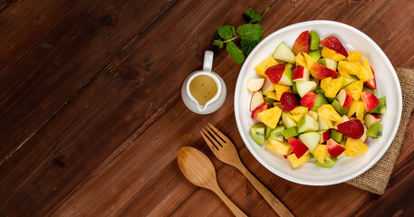 Obraz na płótnie Canvas Mixed fruits salad including strawberry, kiwi, apple, and pineapple in white dish place on sackcloth on wooded table. Utensil and oil salad dressing cup beside with mint leaf decorating